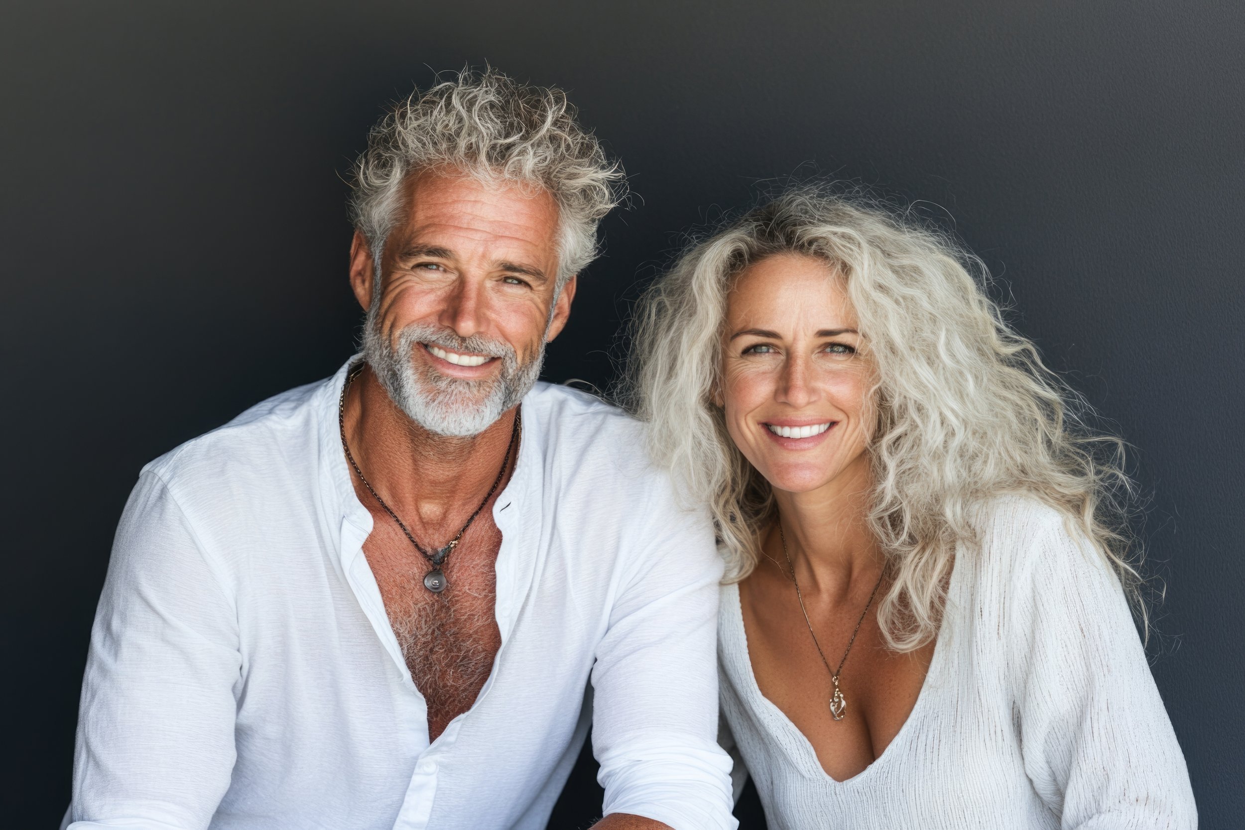 A smiling older man with gray curly hair and beard wearing a white shirt and a necklace, and a smiling woman with long curly blonde hair wearing a white top and a necklace, sitting side by side against a dark background.